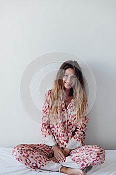 Photo of young happy woman in pajama stretching her arms and smiling while sitting on bed after sleep or nap