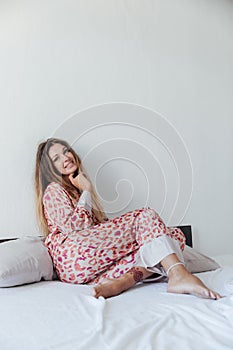 Photo of young happy woman in pajama stretching her arms and smiling while sitting on bed after sleep or nap