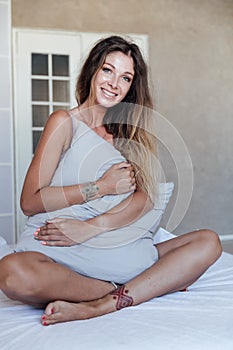 Photo of young happy woman in pajama stretching her arms and smiling while sitting on bed after sleep or nap