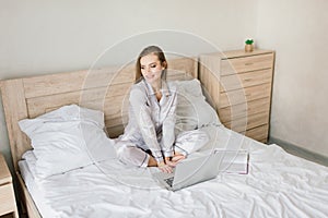 Photo of young joyful woman in pajama with laptop and smiling while sitting on bed in light room