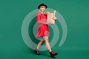 Photo of young lady hold paper bag food walk wear hat red dress footwear isolated green color background