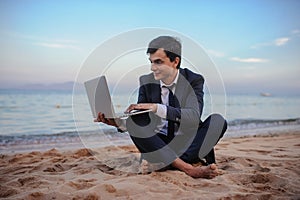 Photo of a young man in suit with laptop working on the beach and talking to someone