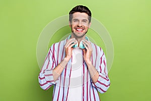 Photo of young optimistic man wearing stylish striped shirt touching his wireless earphones itunes isolated on green photo