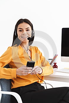 Photo of young phone operator woman wearing microphone headset holding smartphone while working in office