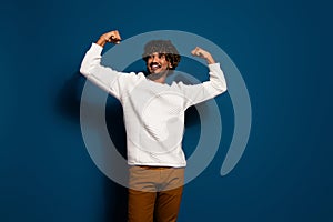 Photo of young sportive guy in white jumper raised fists up showing big biceps triceps muscles isolated over dark blue