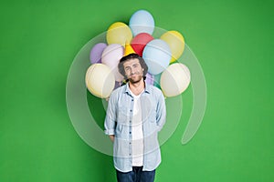 Photo of young stubbled man hold balloons behind back wear paper cone headwear denim shirt jeans isolated green color photo