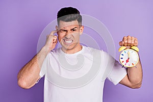 Photo of young unhappy upset man hold hands alarm clock loud noise annoyed isolated on purple color background