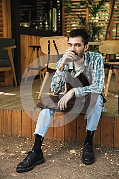 Photo of young waiter man sitting on wooden floor while working in cafe or coffeehouse outdoor