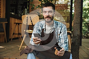 Photo of young waiter man using smartphone while working in cafe or coffeehouse outdoor