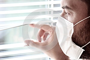 Photo of a young white man with a medical face mask looking out of the window with jalousie during coronavirus quarantine.