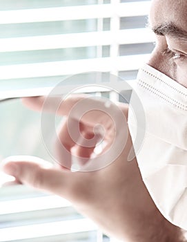 Photo of a young white man with a medical face mask looking out of the window with jalousie during coronavirus quarantine.