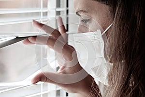 Photo of a young white woman with a medical face mask looking out of the window with jalousie during coronavirus quarantine.