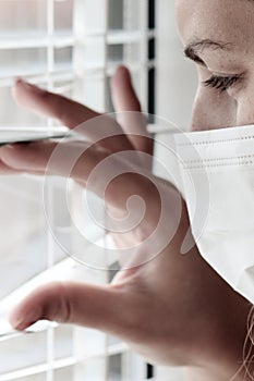 Photo of a young white woman with a medical face mask looking out of the window with jalousie during quarantine.