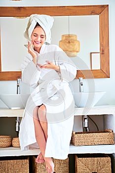 Photo of young woman with bathrobe with cream in her hands standing in bathroom.