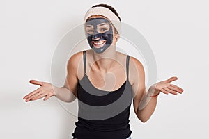 Photo of young woman with charming smile spreading palms aside, shows helpless gesture, wears nourishing facial mask for reducing
