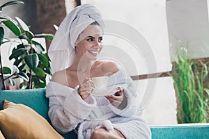 Photo of young woman early morning enjoy coffee spa salon procedure sitting cozy sofa divan wear towels bathrobe indoors