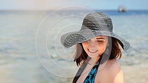 Photo of a young woman in swimsuit and a blue big hat smiling and sunbathing on the beach