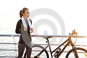 Photo of young woman with towel over neck standing near bicycle outdoors, after workout by ocean