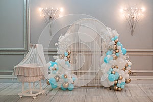 Photo zone of white arc with round part decorated by lights, strings, colorful balloons of different sizes near cradle.