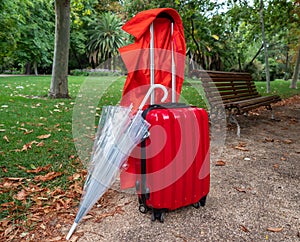 Photograph of a travel suitcase, an umbrella and a red gabardine in a park