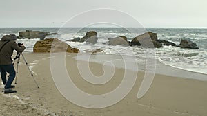 Photographer adjusts camera on tripod on sand beach near rocks and stormy sea.