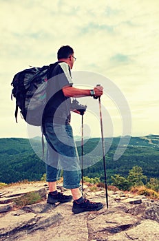 Photographer with big mirror camera on neck and backpack stay on peak of rock. Hilly landscape, fresh green color in valley.