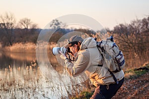 Photographer with camera, telephoto lens and backpack taking picture at lake during sunset