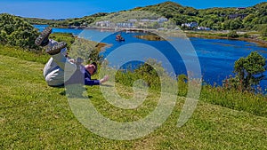 Photographer fallen on the green grass on a hill with the port of Clifden at high tide and a boat in the background