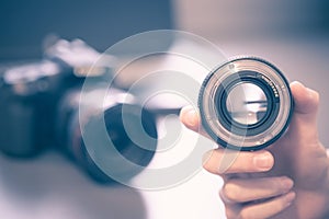 Photographer is holding a photography lens in his hand, camera and laptop in the blurry background