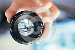 Photographer is holding a photography lens in his hand, laptop in the blurry background