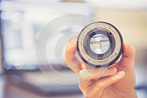 Photographer is holding a photography lens in his hand, laptop in the blurry background