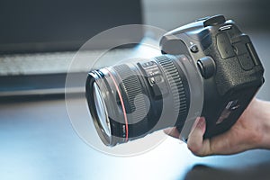 Photographer holds a reflex camera with telephoto lens in his hand. Table and laptop in the blurry background