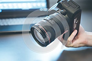 Photographer holds a reflex camera with telephoto lens in his hand. Table and laptop in the blurry background