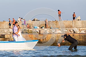 Photographer knee-deep in the water shoots a couple of affectionate newlyweds in a boat in the Black Sea bay, boy is gazing