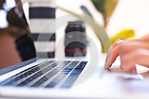 Photographer`s workplace, laptop and camera lenses on a white table, hands on keyboard close up