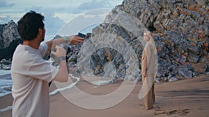 Photographer shooting beach girl in suit. Blonde model posing at rocky seashore