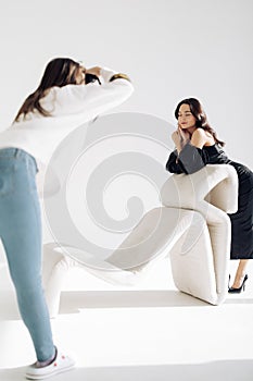 Photographer shoots young woman posing with closed eyes in studio near armchair