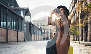 Photographer tourist with suitcase looking on retro photo camera. Smiling girl in hat travels in Barcelona. Sunlight flare street