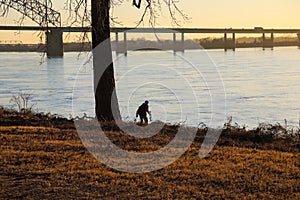 A photographer walking down the banks of the Mississippi river to get a shot at sunset with a bridge over the river