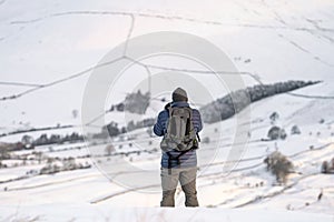 Photographer wrapped up warm in snow covered mountain landscape taking pictures of hillside range with backpack countryside