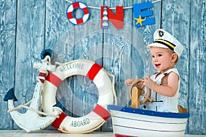 Photoshoot for a boy of one year. Little sea captain, sailor on toy ship with steering wheel. Sea anchor and lifebuoy on gray