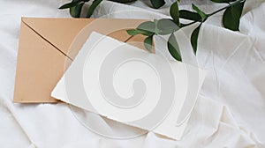 Photostock wedding styled composition. Feminine envelope mockup scene with ruscus leaves, silk ribbon, blank greeting card, on cre