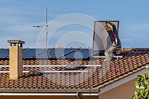 A photovoltaic installer installs a solar panel on a red tiled roof