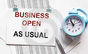 The phrase Business as usual printed on a notebook. Documents, alarm clock and pen lie on a white background photo