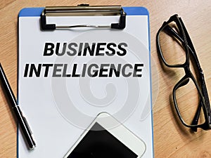 Phrase BUSINESS INTELLIGENCE written on paper clipboard with pen,smartphone and eye glasses.