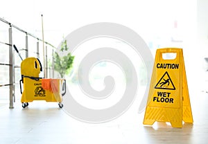 Phrase CAUTION WET FLOOR on safety sign and yellow mop bucket with cleaning supplies, indoors