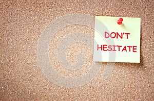 The phrase dont hesitate written on sticky note over cork board