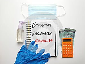 Phrase Economy Recovery Covid-19 on note book with rubber hand glove,sanitizer bottle,mask,pen,calculator and money.