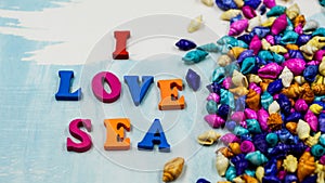 The phrase `I love sea` is composed of colorful wooden letters on a blue and white background and colorful shells.