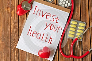 Phrase Invest your health written on sheet of paper, stethoscope and pills on wooden background